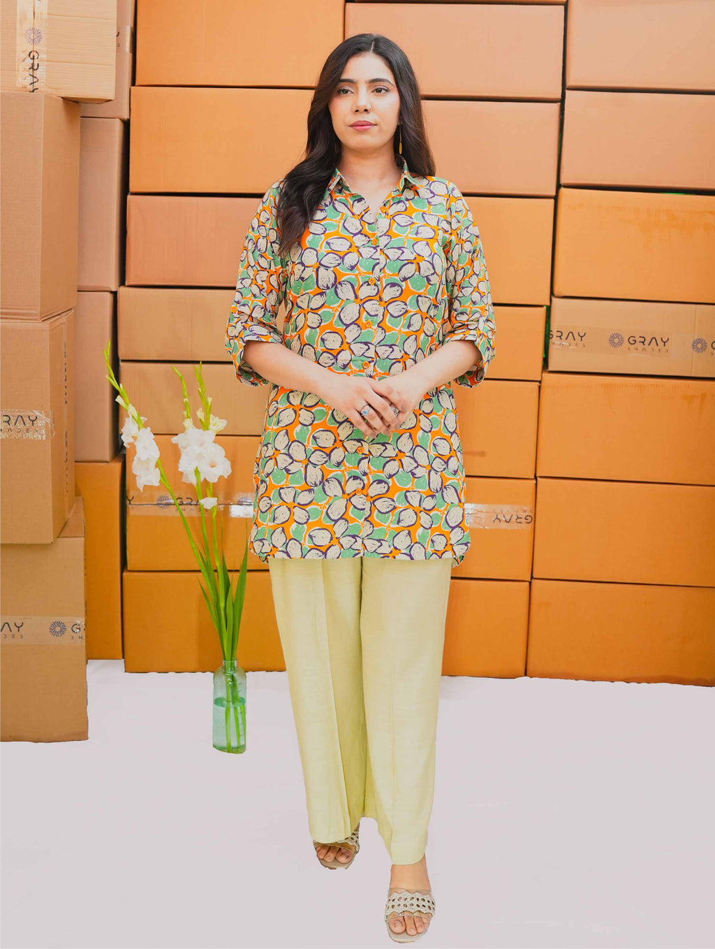 Woman in a patterned top and light pants standing in front of stacked boxes with a vase of flowers.