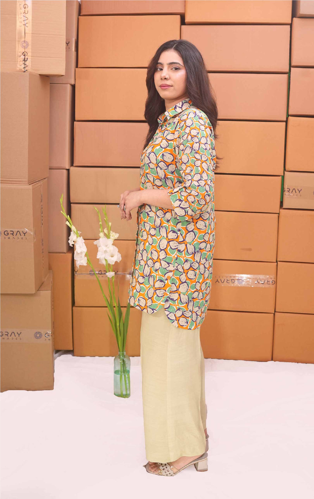 Woman in a patterned top and light pants standing in front of stacked boxes with 'GANT' branding.