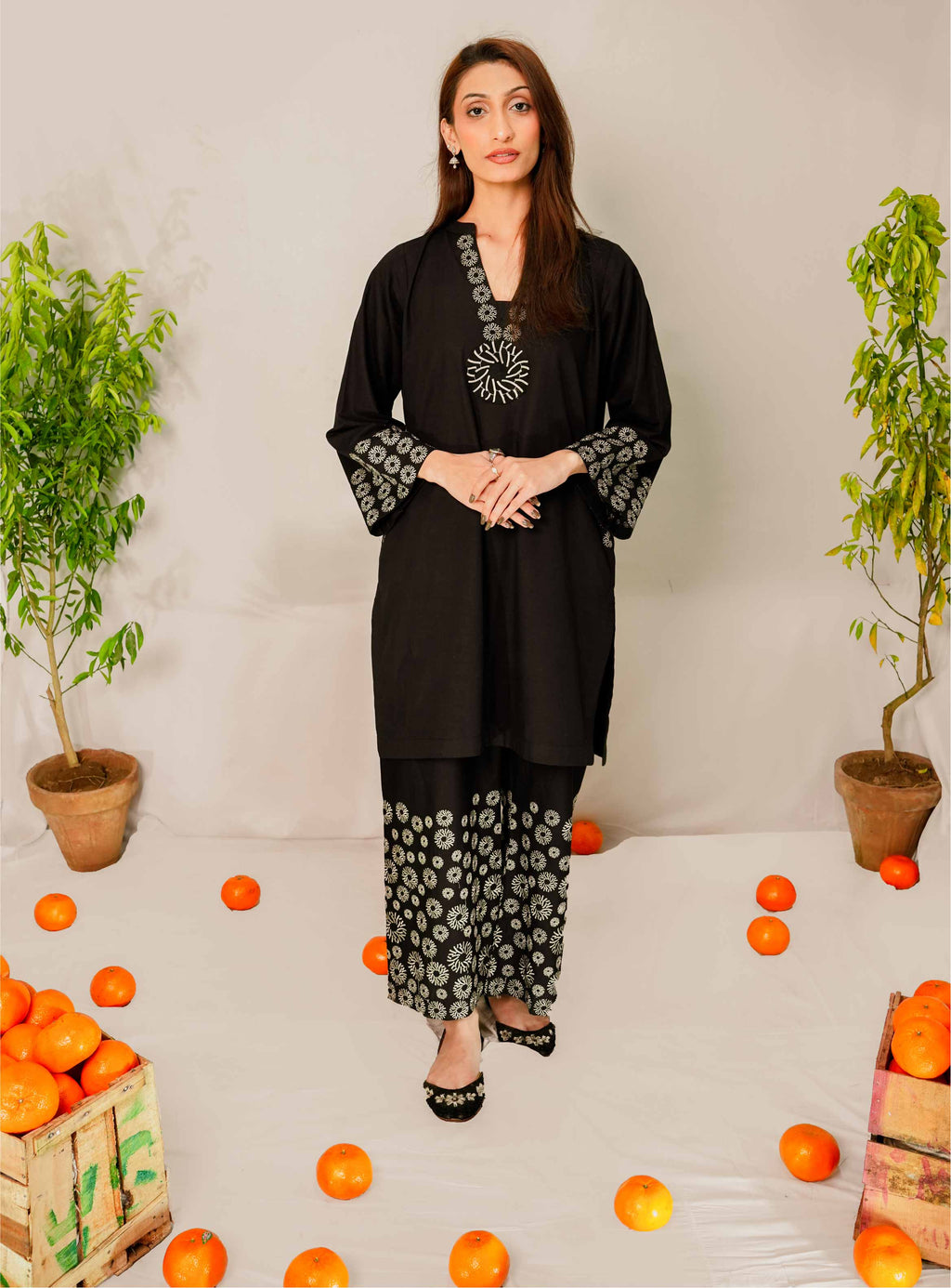 Woman in a black traditional outfit with white patterns standing among oranges and potted plants.
