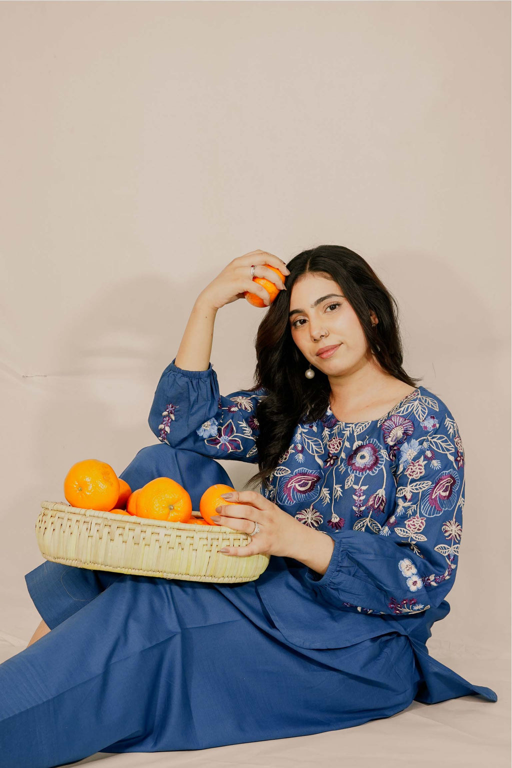 Woman in a blue floral dress holding oranges and a basket against a beige background