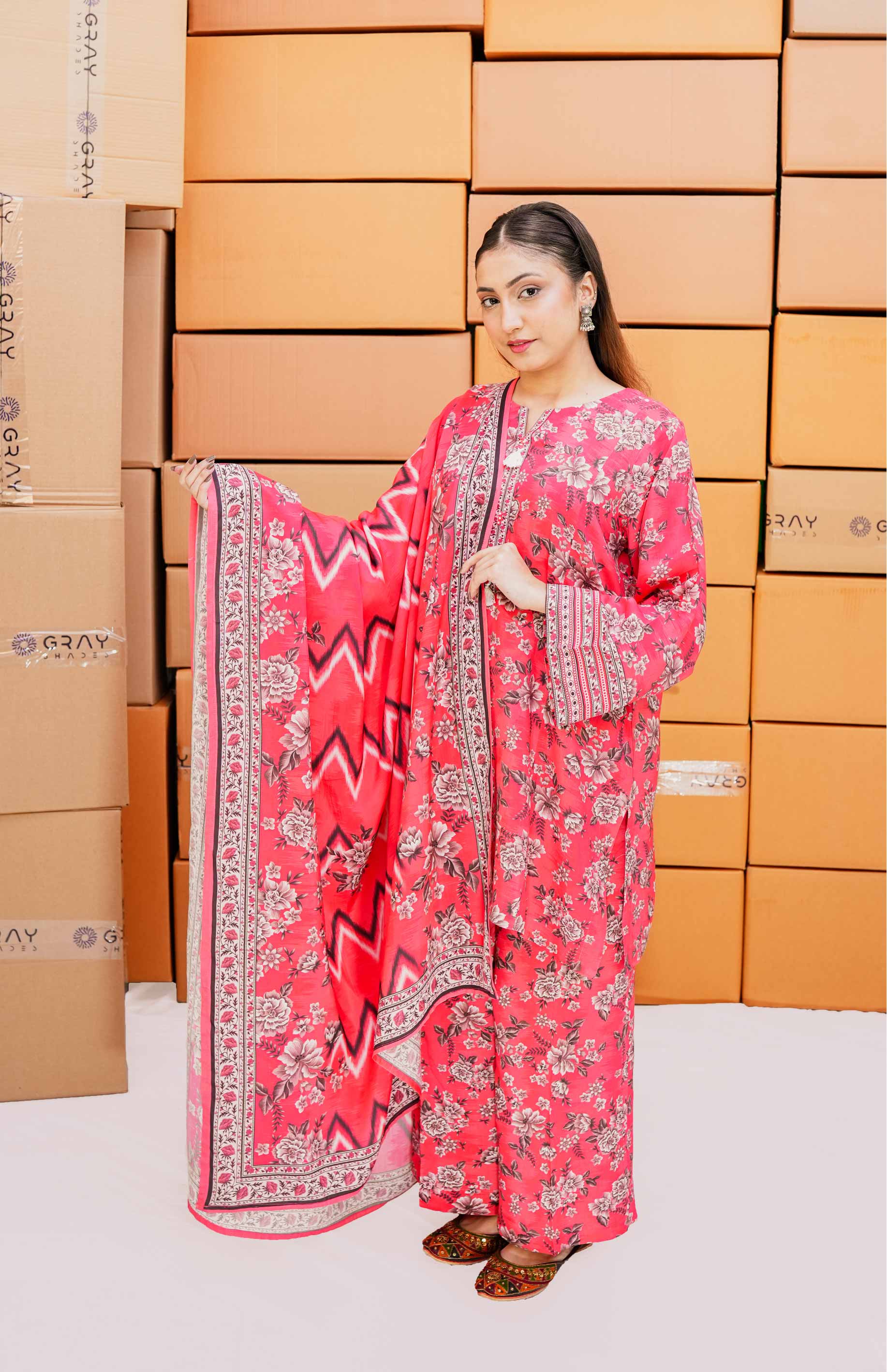 Woman in a pink traditional outfit with a patterned shawl against a background of stacked boxes.