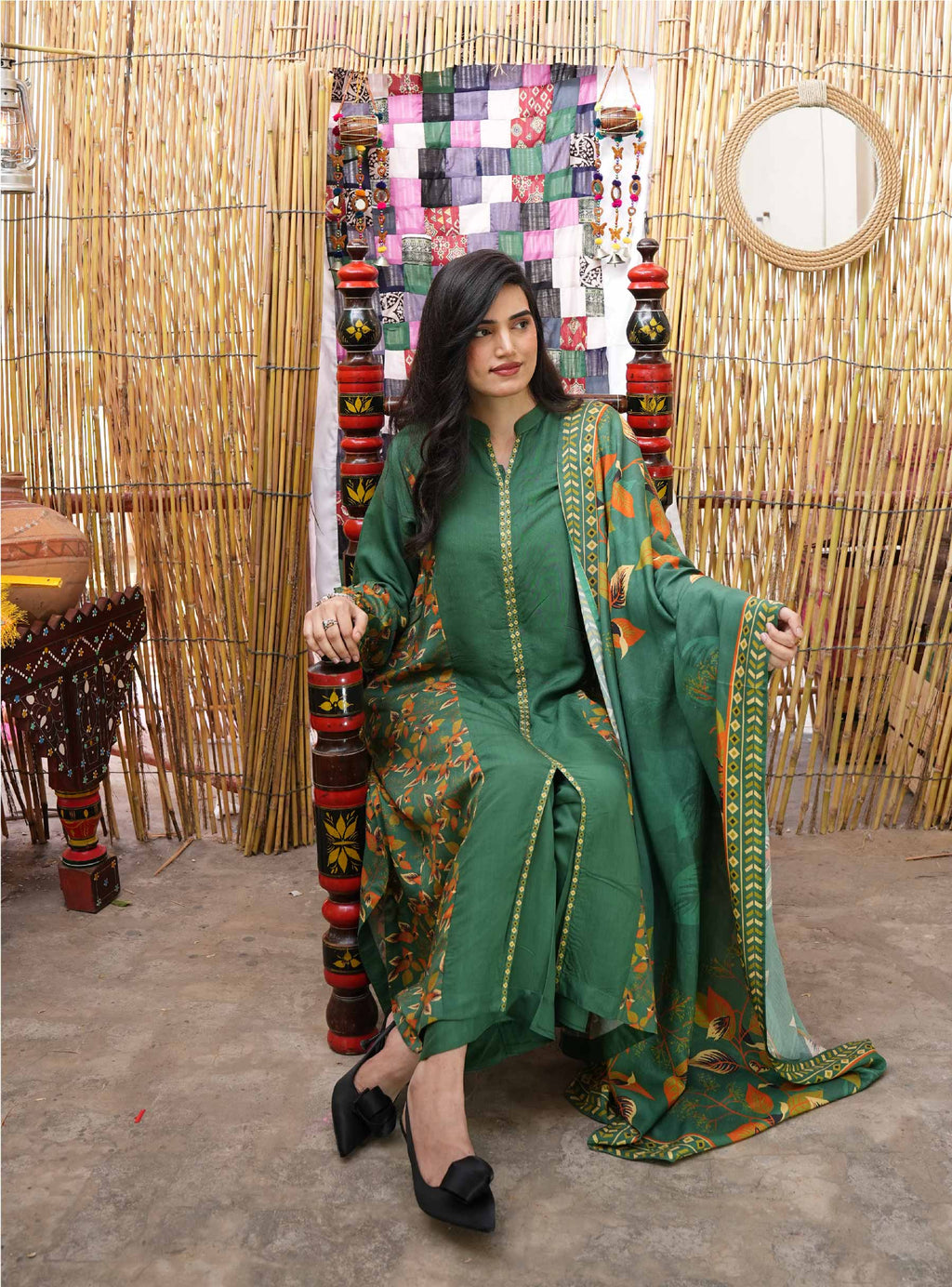 Woman in a green traditional outfit sitting on a decorative chair with a colorful quilt in the background.