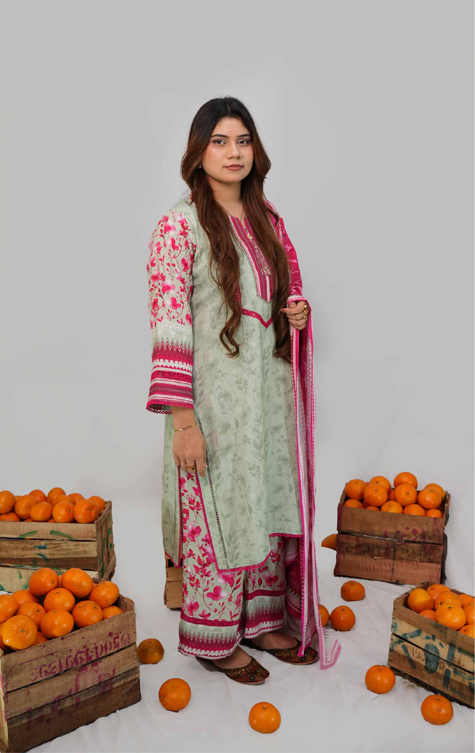 Woman in a traditional outfit standing among crates of oranges on a plain background