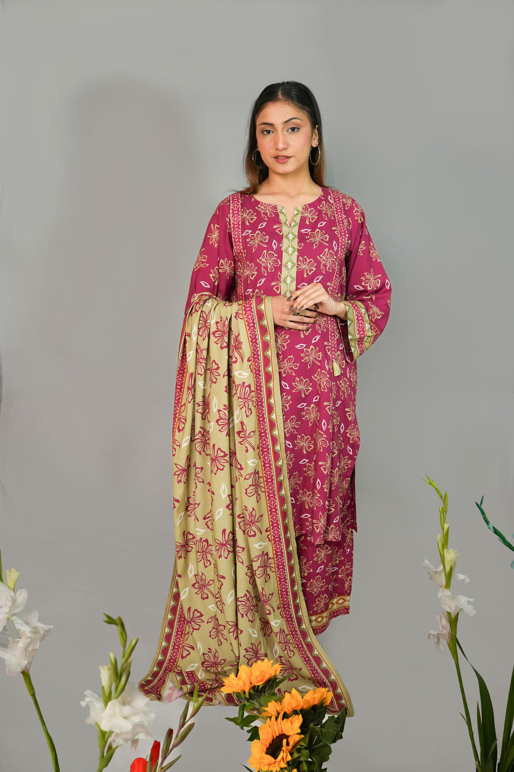 Woman wearing a pink and green traditional outfit with floral patterns, standing against a gray background with flowers in the foreground.