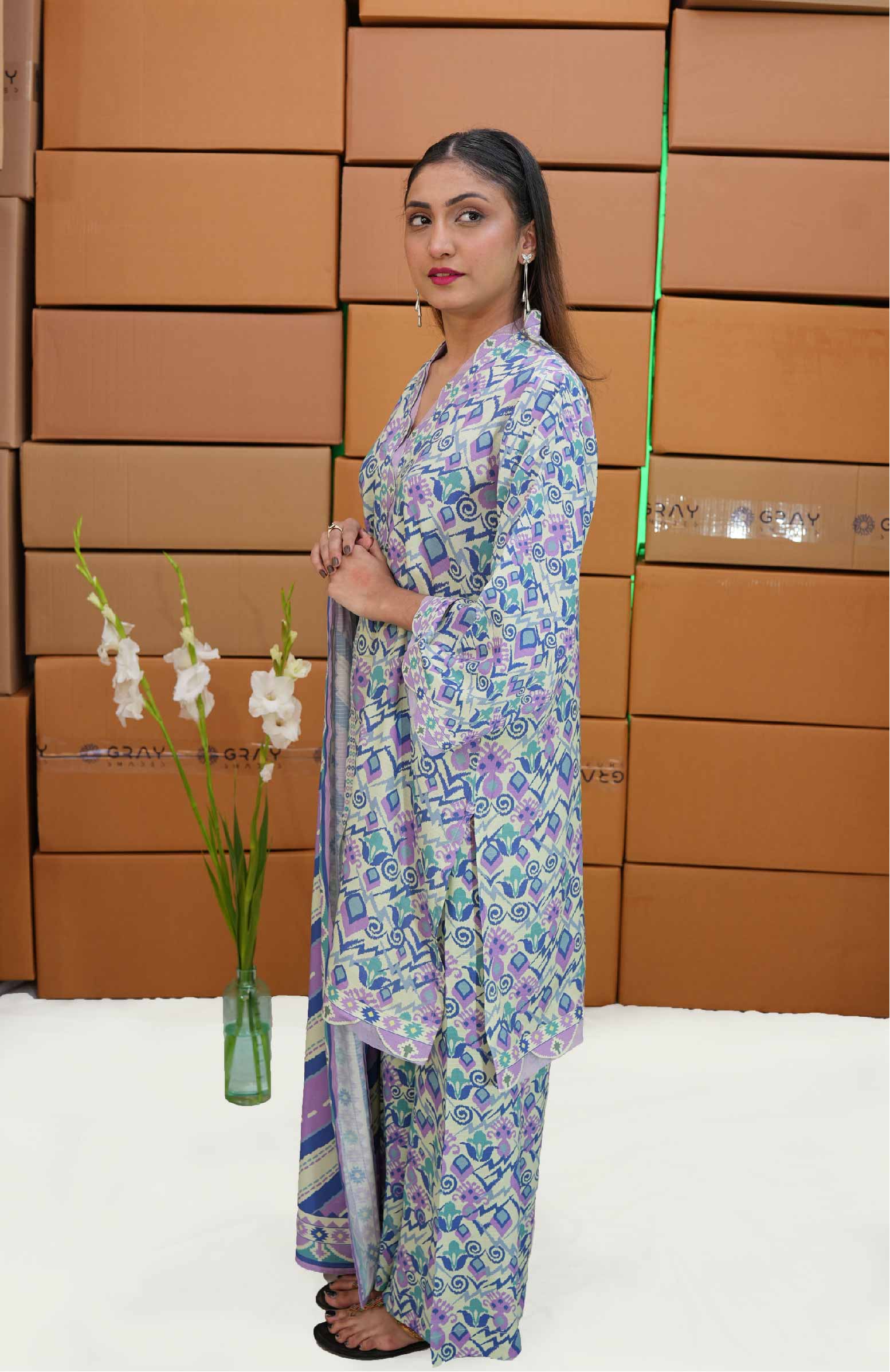 Woman in a floral outfit standing in front of stacked boxes with a vase of flowers on a white floor.