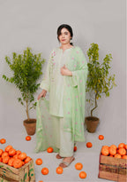 Woman in a light green traditional outfit standing among scattered oranges with plants in the background.